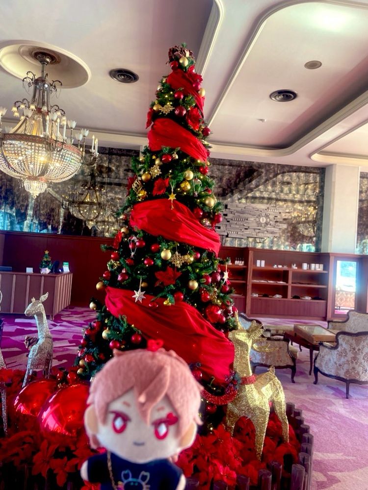 写真。ホテルニューアカオのロビー。大きなクリスマスツリーが飾られている。ツリーの両脇には金と銀のトナカイの置物。天井には豪華なシャンデリア。手前にピントのボケたエスぬい。
