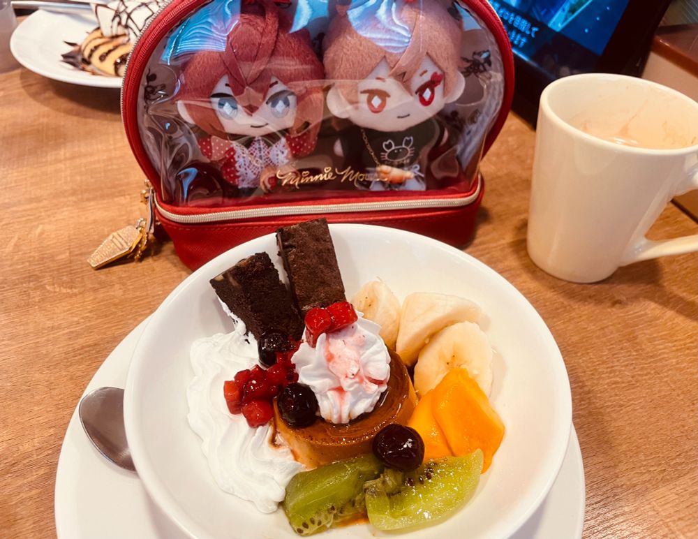 ぬいとファミレスの写真。前面クリアの赤いポーチに入れられたリドルくんとエースのぬいぐるみが、ガストのプリンアラモードを見つめている。プリンの盛り付けの内訳はバナナ、マンゴー、キウイ、ベリーソース、ブラウニー、生クリーム。隣にはコーヒーが入ったカップもある。