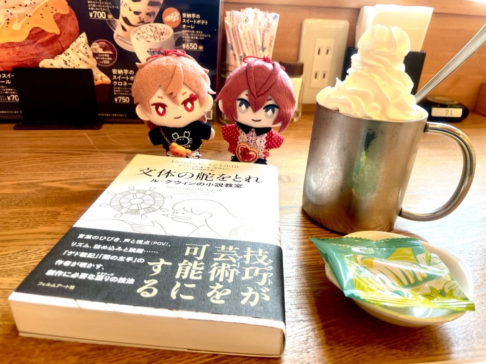 写真。コメダ珈琲店にて。左後方にリドルとエースのぬいぐるみ、左前方に本（『文体の舵をとれ』）、右にアイスウィンナーコーヒーと豆菓子。