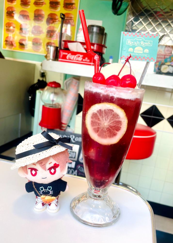 写真。エースのぬいぐるみとクリームソーダ。背景はアメリカンダイナー風の喫茶店。エースのぬいぐるみは麦わら帽子を被り、カニのTシャツを着てカニパンのポシェットを身につけている。チェリー味のクリームソーダの上にはさくらんぼが3つ乗っていて、店のロゴがプリントされた旗がさしてある。