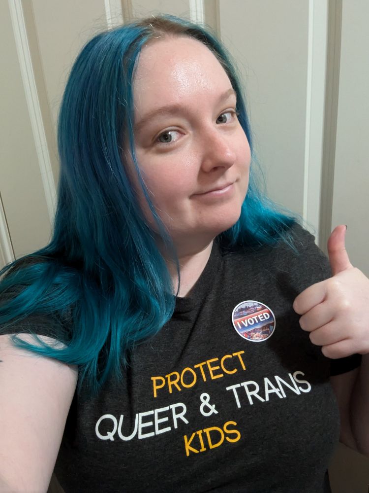 A girl with blue hair poses giving a thumbs-up. Her shirt says "protect queer & trans kids" and has an "I voted" sticker.