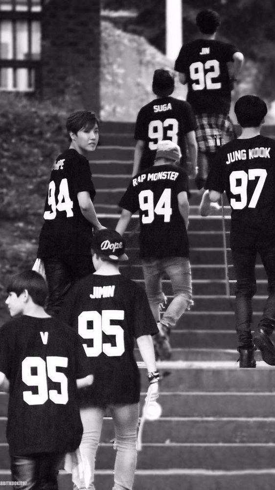 BTS when they were just kids walking up stairts towards a building. All wearing football jerseys with their name and birth year written on the back. Photo is shot from the back, you don’t see there faces except from hobi’s who is looking into the camera.