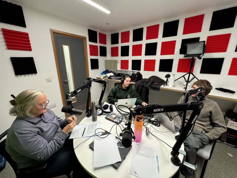 Jan, produce Ffion and guest Darren Axe around the table in the podcast studio, with microphones and production equipment all over the table.