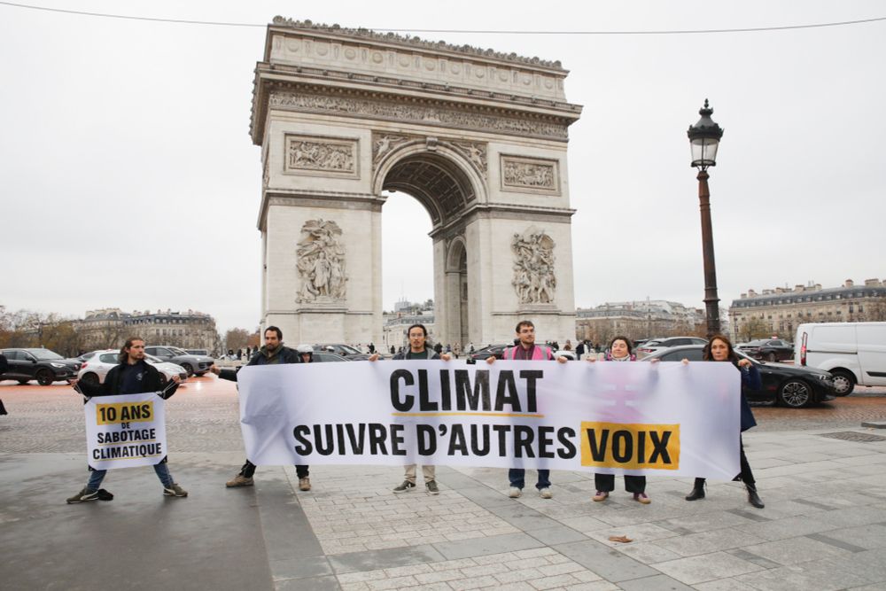 Six activistes tiennent deux banderoles devant l'Arc de Triomphe. Derrière elleux, les voitures répandent de la peinture orange se trouvant sur la chaussée. Sur la grande banderole se trouve le message "CLIMAT - suivre d'autres voix", la petite banderole indique "10 ans de sabotage climatique"