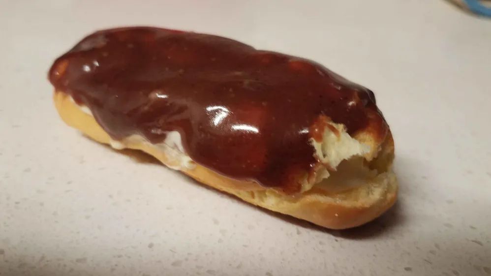 A lone gluten-free eclair sitting on a white granite (?) countertop. The chocolate is glossy and a little vanilla cream filling peeks out the sides.