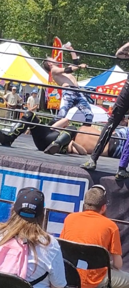 Maxwell standing on the apron of a wrestling ring dumping water directly into his eyes. There is a sitting crowd in the foreground, another man on the apron opposite Maxwell, and two people lying in the ring. The venue is outdoors.