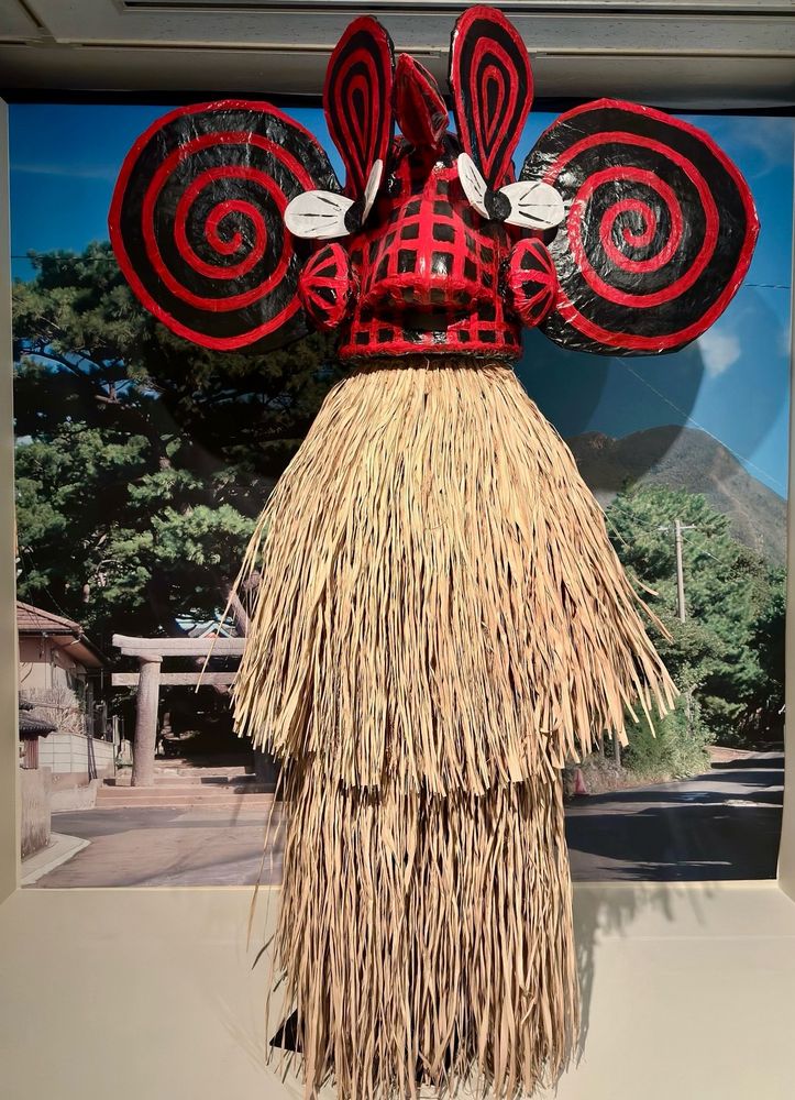 前の画像で説明されたメンドン。真っ赤な顔に白い目。耳の位置には丸く大きな黒いぐるぐる模様がある。背景写真の左側に鳥居が見える熊野神社の境内から現れる。写真左側には今も火山活動が続く硫黄岳がみえる。