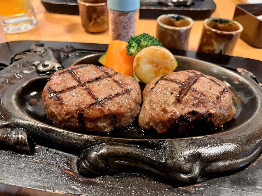 牛を模した鉄板の上で半分に切られたハンバーグが焼かれている。付け合わせのジャガイモ、ブロッコリー、人参、カボチャが乗っている。