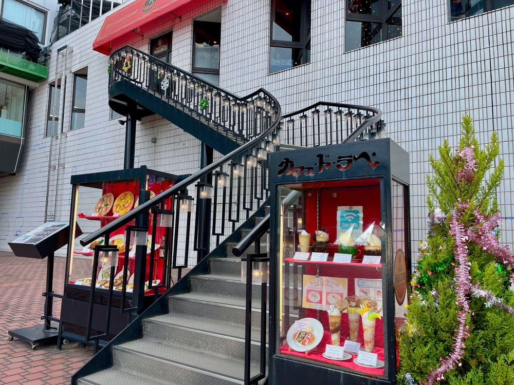 『カフェ・ド・ラペ』さんの外観。食品サンプルの展示ケースが二つ並び、その間から二階に向かって階段が伸びている。階段に点々と付けられた照明は淡く温かな色。右の食品サンプルの横にはクリスマスツリーも飾られている。
