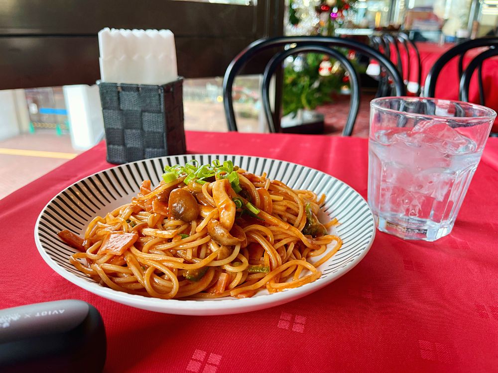 『カフェ・ド・ラペ』さんの窓際の四人がけのテーブル席。赤いテーブルクロスの上にナポリタンと水、紙ナプキンが置かれている。手前には「帰ってきた あぶない刑事」とAVIOTコラボワイヤレスイヤホンのケース。奥にはクリスマスツリーも見える。