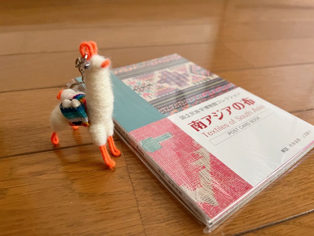 耳と鼻先と足先がオレンジ色、体は白色、背中には卵を入れた荷物を背負ったアルパカのキーホルダーとその向かって右側に「国立民族学博物館コレクション 南アジアの布」ポストカードブックが置かれている。