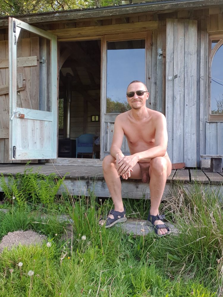 Photo of a naked white man wearing sunglasses sat on the edge of some decking outside a wooden cabin.