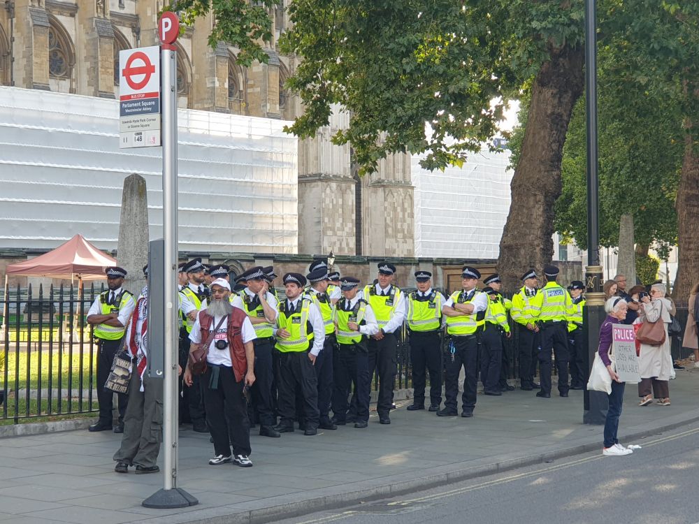 A line of police officers in high viz vests stood next to a bus stop, as if they're waiting for a bus.
