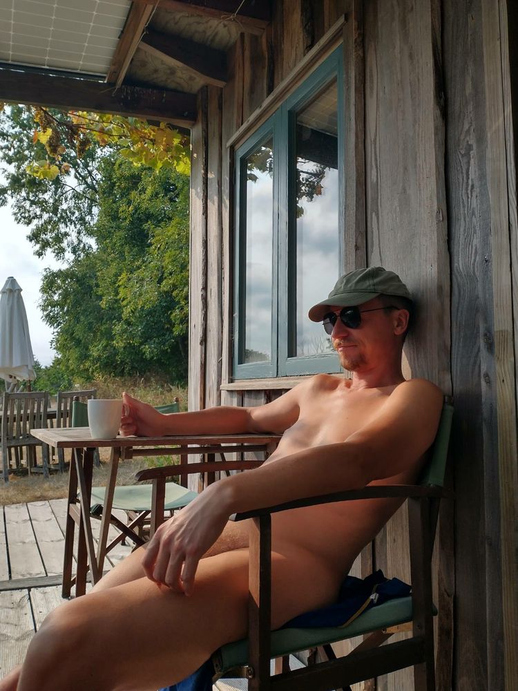 Photo of me, a white 40-something man, sat naked on a camping chair outside a wooden cabin. I'm wearing sunglasses and a dark green cap. I'm holding a white mug in one hand.