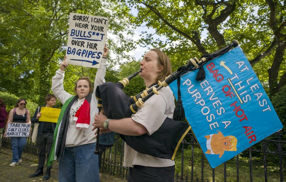 Two protesters, the one on the right blowing into bagpipes, holding signs. One on teh right reads "At least THIS bag of hot air serves a purpose", indicatinng the Bagpipes. The protestor on the left. hold a sign pointing to the bagpipes, reading "Sorry, I can't hear your bullsh**t over her Bagpipes"