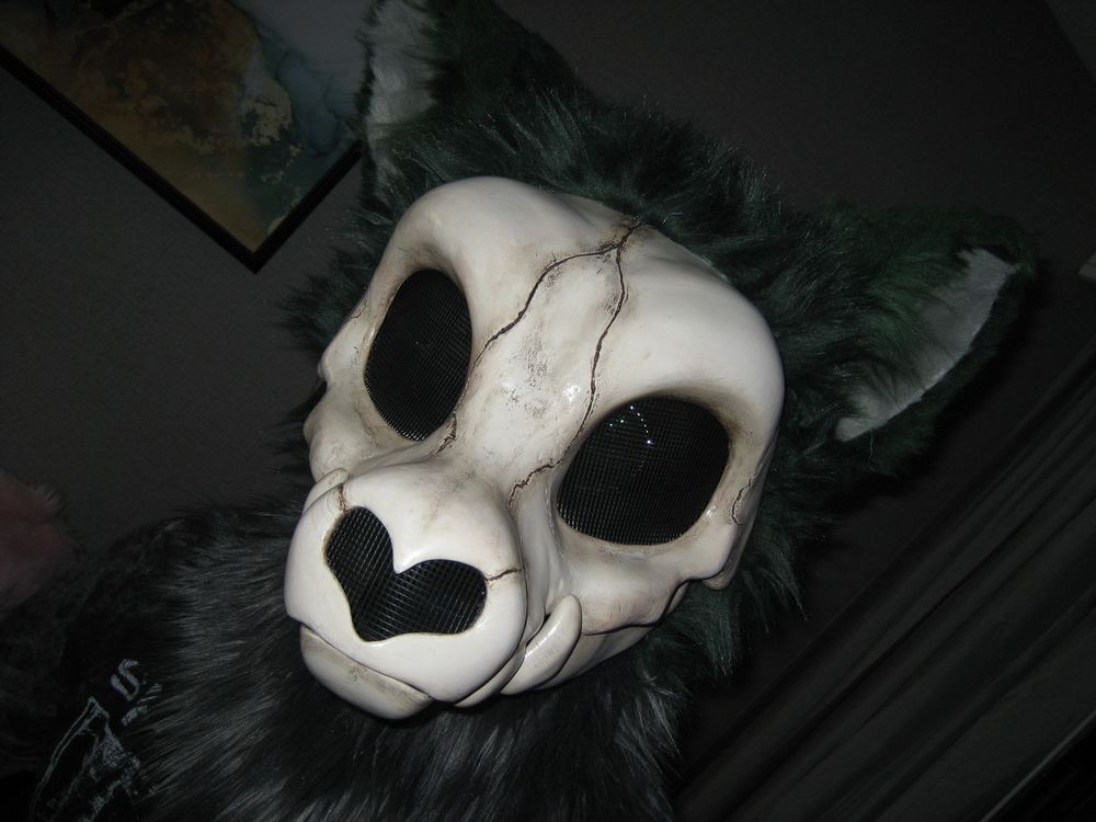 digital flash photo of a skull-creature fursuiter in a dark hotel room; the skull has dark cracks running through it, and the fur around the back of its head and ears is dark green; the inner ears are light gray, and the neck floof is a darker gray color