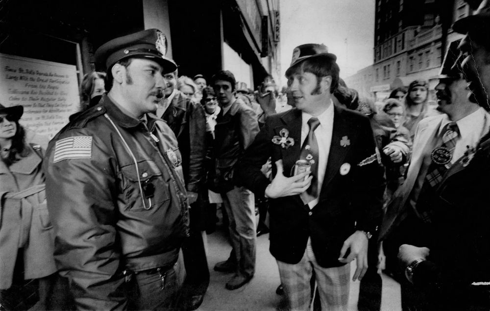 A police officer gives a stern look to a man dressed in St. Patrick’s Day regalia who is carrying a beer on the sidewalk. 
