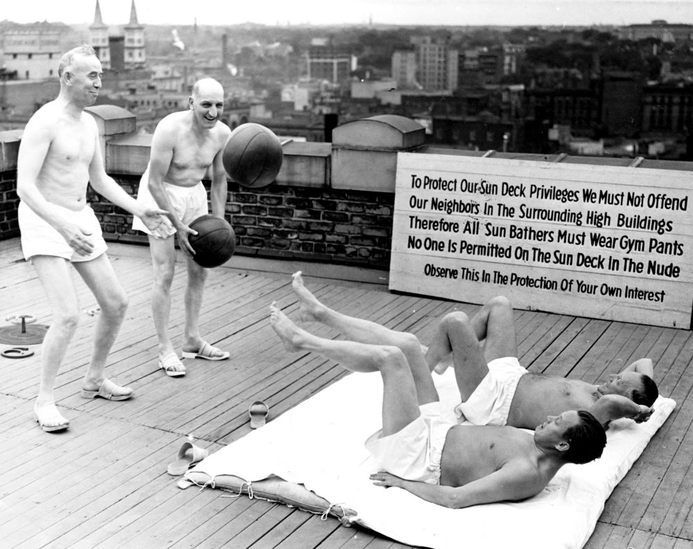 Four men in gym shorts and sandals exercise with medicine balls on the roof of a building. A sign next to them reads, “To protect our sun deck privileges, we must not offend our neighbors in the surrounding tall buildings. Therefore all sun bathers must wear gym pants. No one is permitted on the sun deck in the nude.”