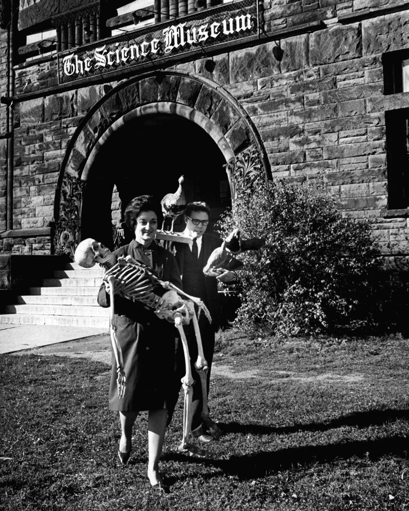 A woman carries a skeleton out of a building with a sign above the door that reads “The Science Museum,” while a man beside her carries taxidermy animals. 