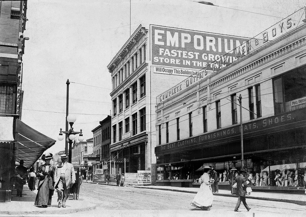 A pair of pedestrians walk down a sidewalk while two others cross a street in a busy downtown street scene from 1906. Behind them, a building under construction has a sign painted on its wall that reads, “Emporium: Fastest growing store in the Twin Cities.”