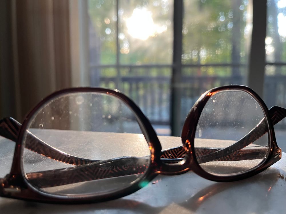 A pair of reading glasses upside down on a white surface with sunshine coming in from behind lighting up the lenses.