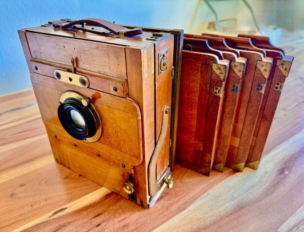 Ein antiker, hölzerner Großformat-Fotoapparat steht auf einem Holztisch.

Der Apparat ist aus hellem Holz gefertigt und mit Metalldetails und -elementen verziert.  Hinter der Kamera stehen vier Holzplatten.