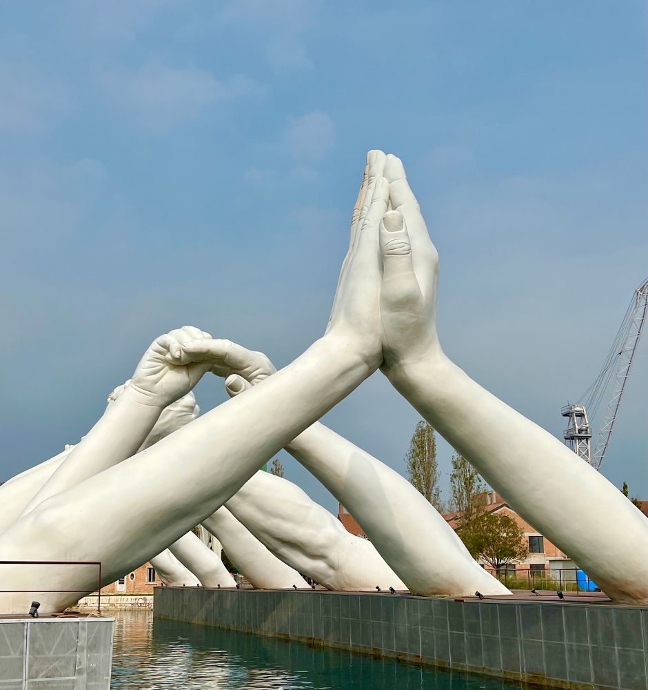 Die Szene zeigt mehrere große, weiße Skulpturen von Armen und Händen, die sich über einem Wasserbecken erstrecken und miteinander verflochten sind. Die Hände sind in verschiedenen Haltungen angeordnet, die eine starke symbolische Kraft hervorrufen. Die Arme sind realistisch modelliert und in verschiedenen Positionen angeordnet