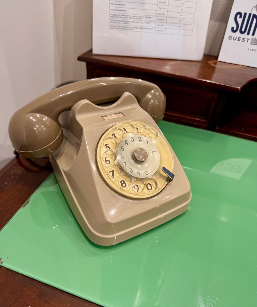 Ein altes grau/beiges Telefon mit Wählscheibe steht auf einer Holzplatte mit grüner Abdeckung. Auf der Wählscheibe steht klein: Italtel.