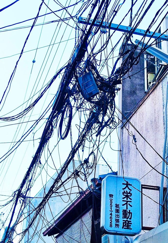Extremes Kabelgewirr an einer Hauswand über hellem Himmel. An einem Haus hängt ein Schild mit japanischen Zeichen und einigen Zahlen.,