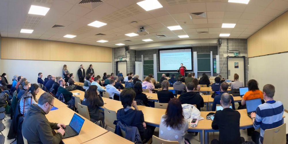 Panorama of the room during a talk, taken from the back. It’s a classroom with multiple rows of tables and seats. Some people are standing on the side. The speaker is in front of their slides at the front of the room.