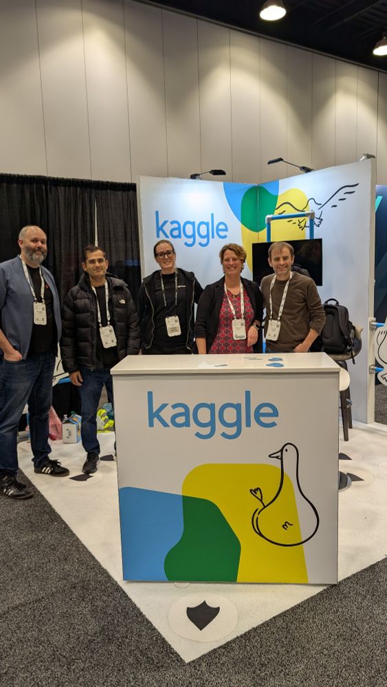 Five people standing in front of a bright and cool Kaggle booth.