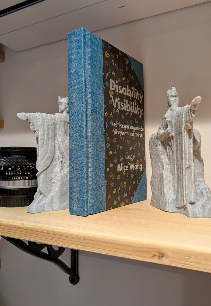 A custom-bound copy of Disability Visibility by Alice Wong stands upright on a wooden shelf between two grey stone-textured 3D-printed statues of the The Pillars of the Kings from Lord of the Rings. The spine of the book, covered in textured denim bookcloth, is titled in silver foil. The front board features disability-themed icons printed on black paper with the icons in the colours found on the disability pride flag.