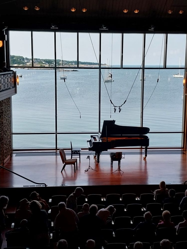 photo of a piano and a couple of chairs and music stands on a stage, in front of a big window looking out over the ocean