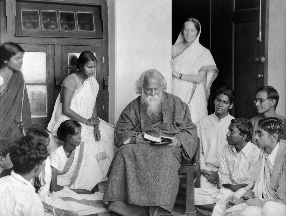 black and white photo of an older man with white hair and beard in a dark robe holding a book on a veranda with men and women and women in South Asian attire gathered around him listening and smiling 