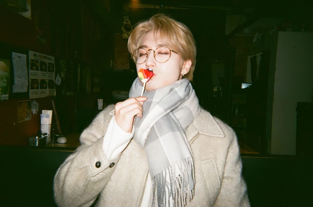 Blond HAN in a beige fuzzy winter coat and thick gray scarf wrapped around his neck. He is wearing round, wire-rimmed glasses and kissing a red heart-shaped lollipop. 