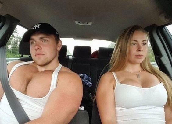 A man and woman sit next to each other in the front of a car, each of their voluminous chests threatening to burst from their tank tops.