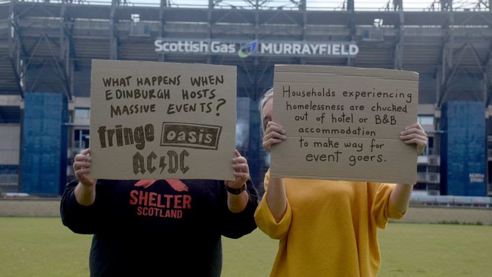 A photograph or two people standing in front of Edinburgh's Murrayfield Stadium, holding cardboard signs. The person on the left's sign says "What happens when Edinburgh hosts massive events?" with logos for Edinburgh Fringe, Oasis & AC/DC underneath.

The person the right's sign says "Households experiencing homelessness are chucked out of hotel or B&B accommodation to make way for event goers"