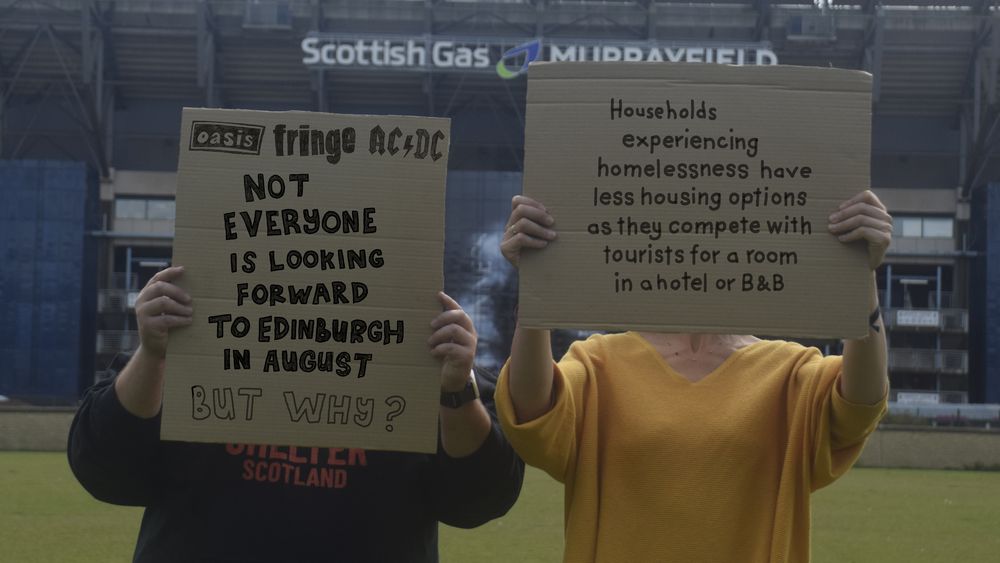 Two people holding signs up outside of Murrayfield stadium in Edinburgh. One sign says: Not everyone is looking forward to Edinburgh in August. Why why?", and the other sign says: "Households experiencing homelessness have less housing options as they compete with tourists for a room in a hotel or B&B"