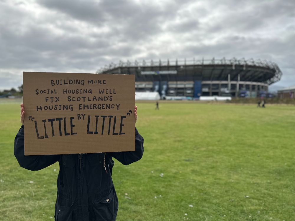 Someone holding a sign outside of Murrayfield Stadium, where Oasis will be playing this Friday, Saturday and Tuesday, that says: "Building more social housing will fix Scotland's housing emergency "Little by Little" (in reference to one of Oasis's songs)