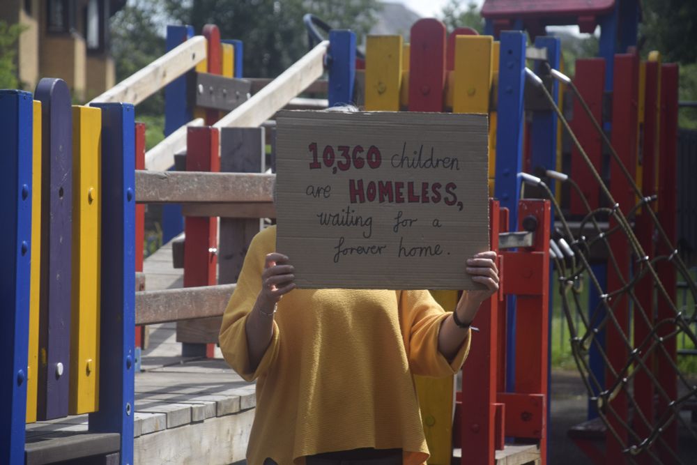 A woman standing in an empty playground. She's holding a large cardboard sign that says "10,360 children are homeless, waiting for a forever home"