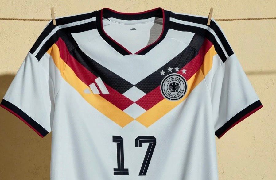 Das von Adidas vorgestellte Trikot für die Fußball-WM 2026: Es ist weiß mit den typischen schwarzen Adidas-Streifen auf den Schultern und einem dünnen schwarzgeränderten  Halsausschnitt. Von beiden Seiten verlaufen diagonale schwarz-rot-goldene Streifen zur Mitte, wo sie sich angeschrägt treffen. Es ähnelt dem Weltmeistertrikot von 1990.