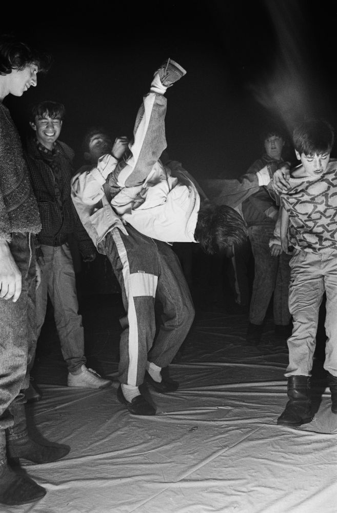 Foto aus einer Dorfdisco in den 1980er Jahren. In der Mitte steht ein Jugendlicher, der einen anderen am Bein haltend durch die Luft wirbelt. Der Boden ist mit Plastikfolie ausgelegt und ein weiterer Jugendlicher tanzt in einem Strickpullover, während andere über die Szene lachen.