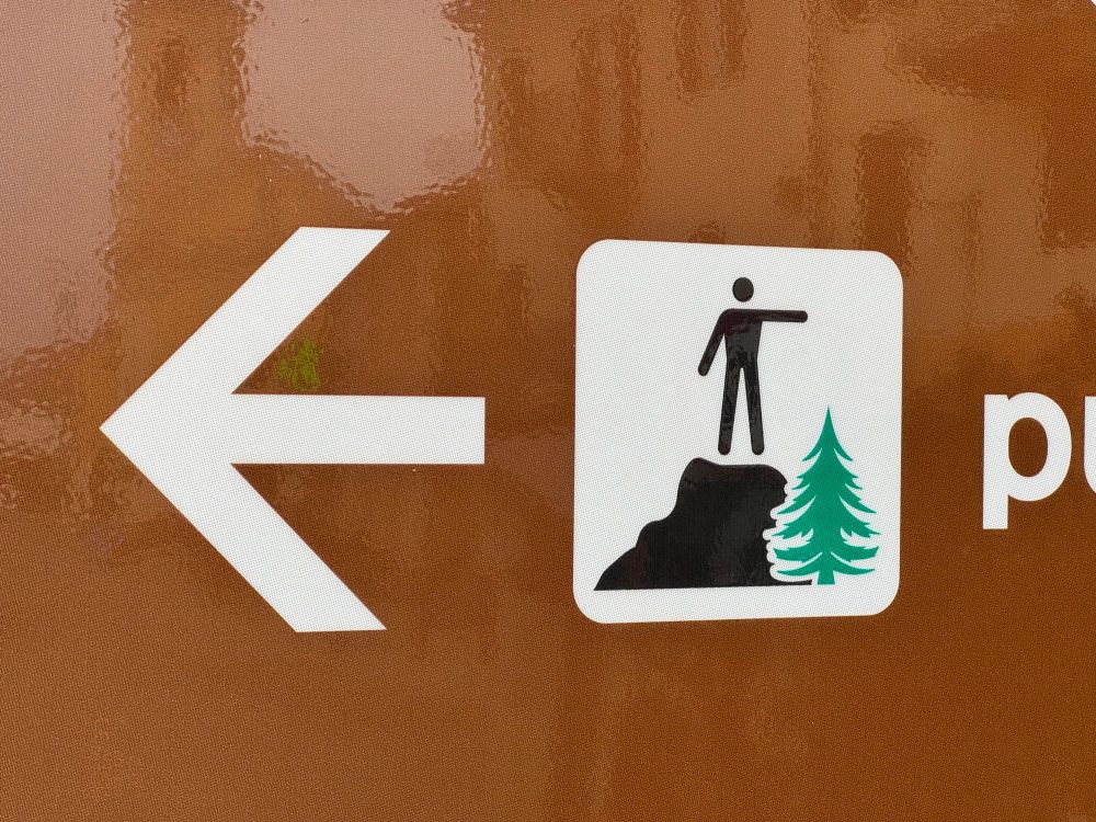 Ein großer Pfeil und links daneben ein Piktogramm, das einen Hügel zeigt, auf dem eine Person mit ausgestreckten Arm steht. Unterhalb des Arms ist eine grüne Tanne