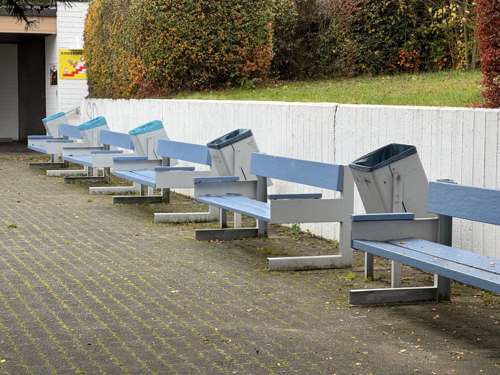 Fünf hellblaue Sitzbänke zwischen denen leicht geneigte Mülleimer montiert sind.