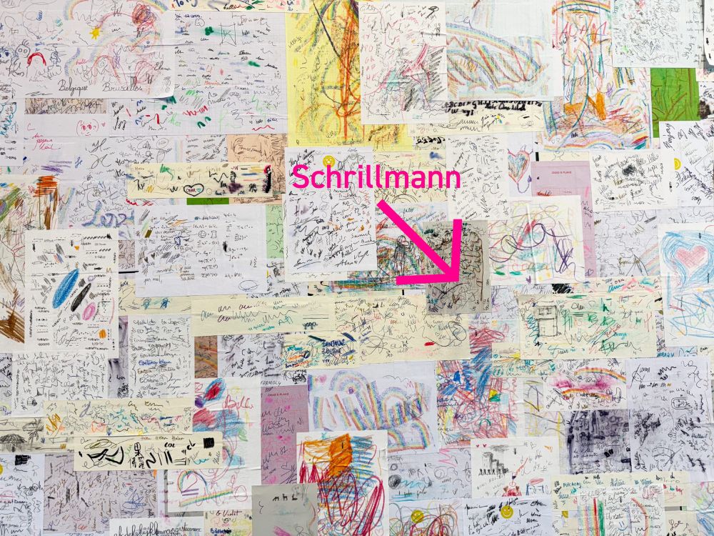Eine ganze Wand mit bekritzelten Papierzetteln zum Ausprobieren von Stiften in einem Schreibwarenladen. Ein Pfeil und der Name Schrillmann zeigen in das Gekritzel.