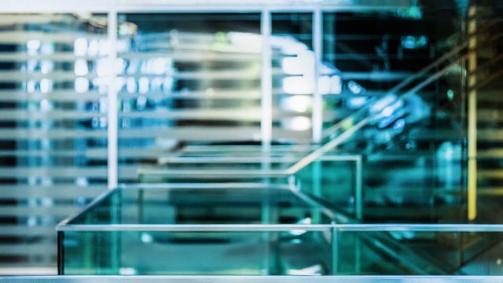 Grünlich-blaues Foto eines Treppenhauses mit viel Glas, Metallhandläufen und einem Hintergrund mit unscharfen horizontalen Lamellen. Es ist der Bildhintergrund, vor dem das häufig genutzte Foto des Österreichers Stefan Weber fotografiert wurde, der u.a. für das Portal NIUS arbeitet.