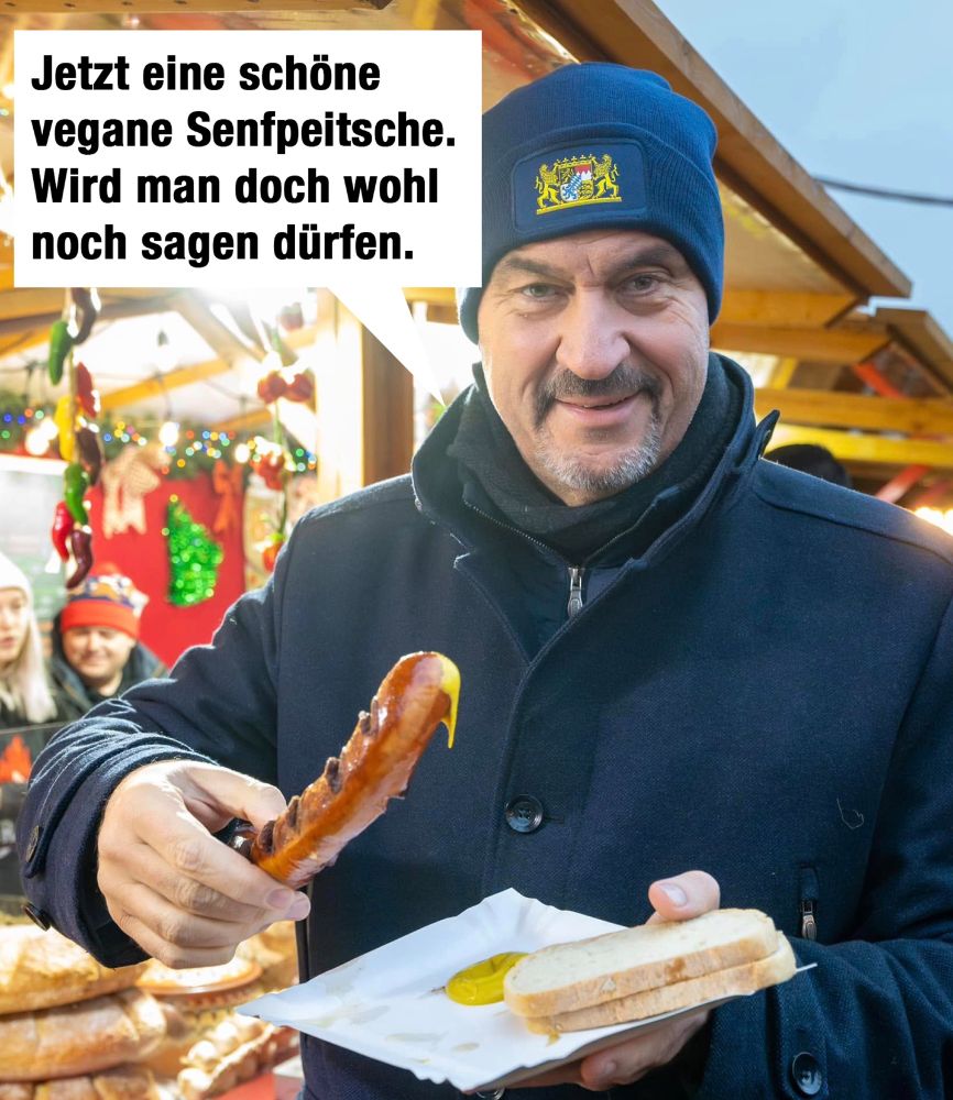 Foto von Markus Söder auf einem Weihnachtsmarkt. Er trägt eine blaue Winterjacke und eine bescheuerte Mütze mit dem bayerischen Staatswappen. In der rechten Hand hat er eine große Wurst, von der der Senf heruntertrieft. Über ihm ist eine Sprechblase mit dem Text: "Jetzt eine schöne vegane Senfpeitsche. Wird man doch wohl noch sagen dürfen.“