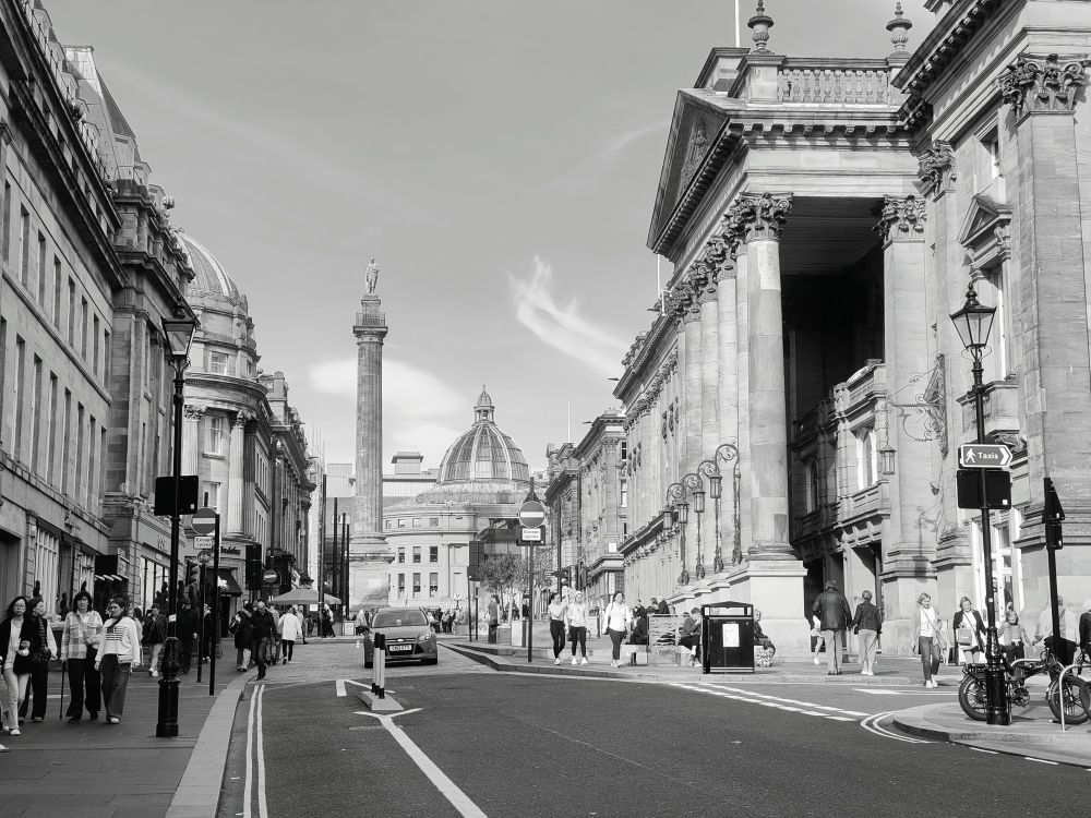 This black and white image shows Grey Street on a sunny October day in Newcastle upon Tyne, England, with Grey's Monument and the Theatre Royal visible in the background.
Grey Street is a historic commercial street in the city centre of Newcastle upon Tyne, known for its Georgian architecture. 
The street is part of the Grainger Town area, which was developed by Richard Grainger between 1824 and 1841. It has more Georgian buildings than anywhere outside London and Bath.

Grey's Monument is a Grade I listed monument built in 1838 to honour Charles Grey, 2nd Earl Grey, who served as Prime Minister and passed the Great Reform Act of 1832.

It has a total height of 133 ft (41 m). It was funded via public subscription and consists of a statue of Earl Grey on a pedestal standing on top of a Roman Doric column. The column was designed by local architect, Benjamin Green, and the statue was created by the sculptor, Edward Hodges Baily.

The Theatre Royal is a Grade I listed building on Grey Street that was designed by architects John and Benjamin Green