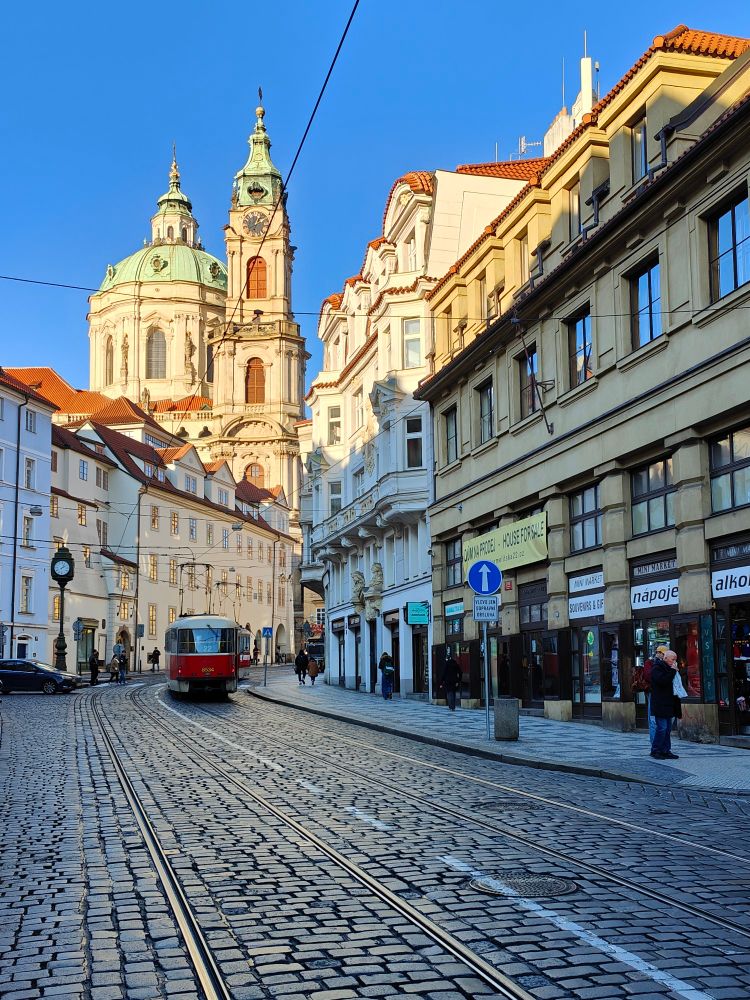 This image shows a street view on a glorious November day of Karmelitská street in the Malá Strana (Lesser Town) district of Prague, Czech Republic.
The prominent green dome and bell tower of the Baroque-style Church of Saint Nicholas is in the background. 
The street is paved with traditional cobblestones and features tram tracks running down the centre, with a red and white tram visible. 
The architecture is typically Baroque, characterized by historic buildings with pastel coloured façades and red-tiled  roofs.
The sky behind the view is bright blue and enhances the creamy hues of the church and some of the pastel coloured buildings not in shade.
The Church of Saint Nicholas (Kostel svatého Mikuláše) is a well-known landmark and one of the most significant Baroque buildings in Prague.