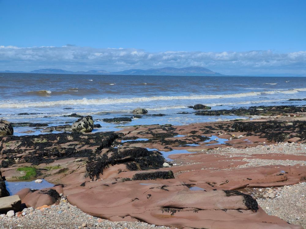 A view from England on a sunny April day across the Solway Firth to Scotland.
In the foreground is a bed of flat red sandstone rock, some of it covered in seaweed. There are some patches of shingle around the rocks.
The sea behind this is slightly choppy with a line of waves breaking near the land.
In the distance is land in the shape of rolling hills, the highest is Criffel.
The sky is blue with a line of clouds just above the hills.